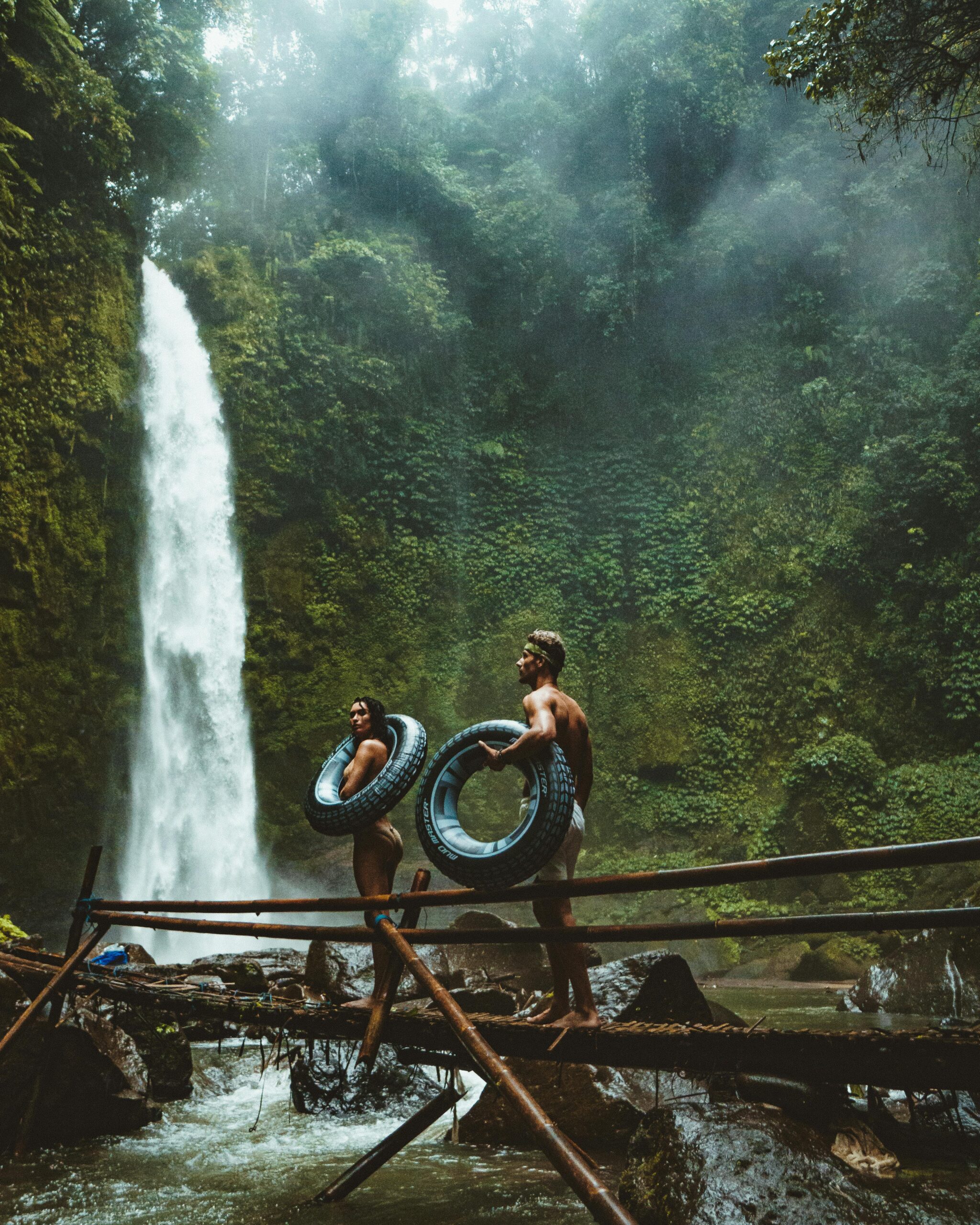 A couple in swimwear with float tubes exploring a lush waterfall in Bali, Indonesia.